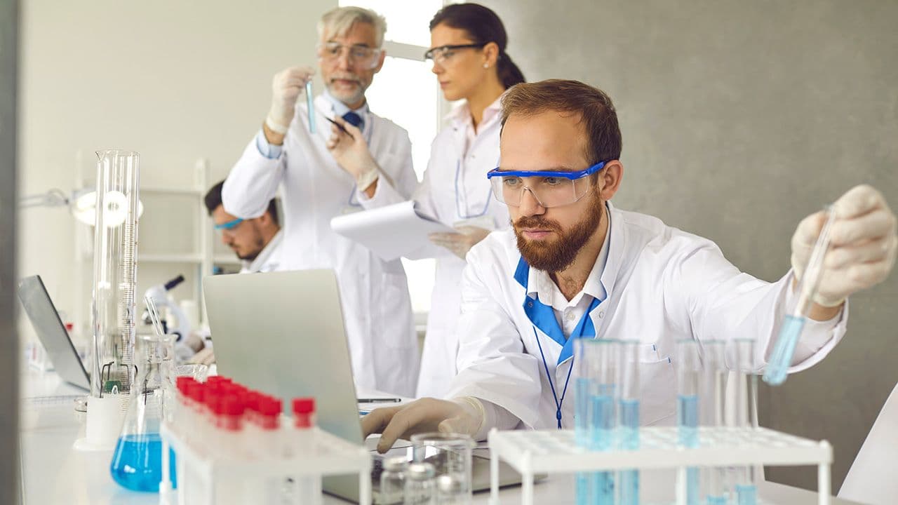 Focused male scientist in protective goggles doing medical research in laboratory, working with lab samples in test glass tubes and entering data in electronic online database on laptop computer | Image Credit: © Studio Romantic - © Studio Romantic – stock.adobe.com